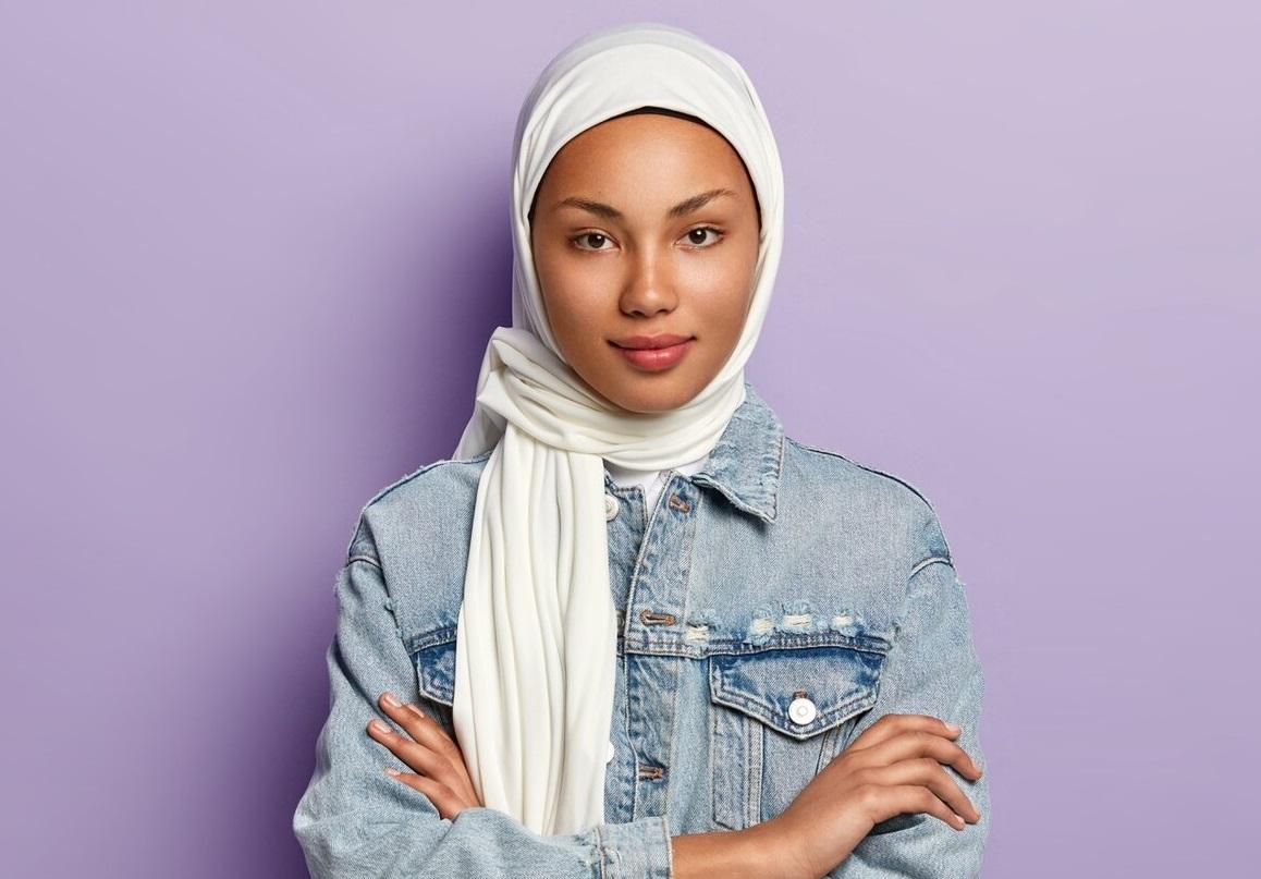 attractive eastern woman covers head with white headscarf guard her dignity power has special dress code keeps hands crossed looks with modesty poses purple wall islamic rules 273609 26323