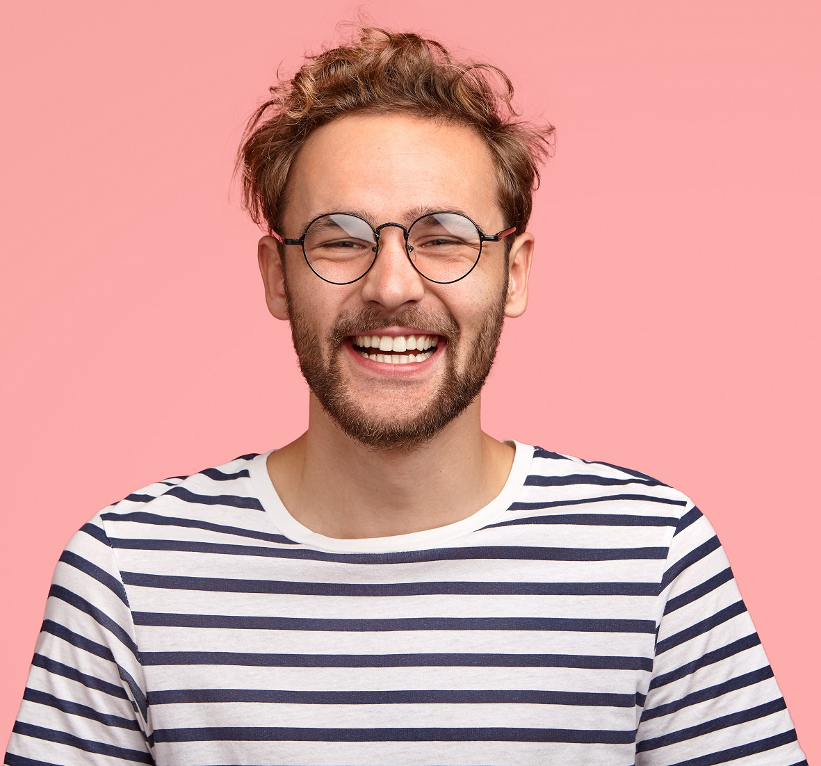 headshot of pleased hipster has satisfied expression, curly hair and bristle, wears round transparent glasses and striped t shirt, feels glad after promotion at work, isolated over pink background